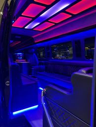 Luxury limousine/party-van interior with quilted leather seating, captain’s chair and bar console, glowing blue and magenta LED ceiling panels and floor strips, windows reflecting exterior lights.