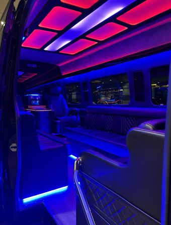 Luxury limousine/party-van interior with quilted leather seating, captain’s chair and bar console, glowing blue and magenta LED ceiling panels and floor strips, windows reflecting exterior lights.