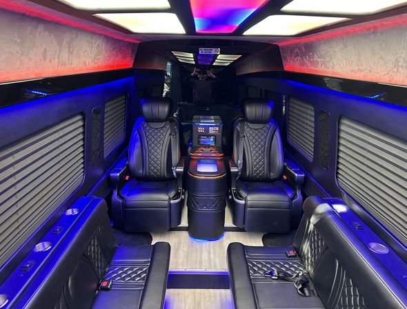 Luxury passenger van interior with black quilted leather captain seats, central console, blue and purple LED ambient lighting and window blinds — executive shuttle vibe.