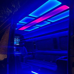 Luxury van interior at night with glowing blue and magenta LED ceiling panels, quilted leather bench seating, captain's chair and touchscreen entertainment console