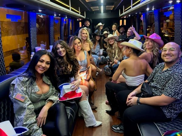 Smiling group of friends inside a limo-style party bus, wearing cowboy hats and casual party outfits, holding drinks and small disco balls under blue LED lighting for a festive nightlife celebration.