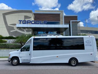 White shuttle bus parked outside a modern golf-entertainment venue with palm trees, glass façade and a bright blue sky with clouds.