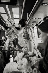Bride and bridesmaids laughing inside a party bus, holding bouquets, a beer bottle and red cup during a wedding day celebration