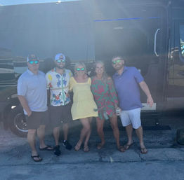 Five friends in colorful summer outfits and mirrored sunglasses posing in front of a black party van in a sunny parking lot