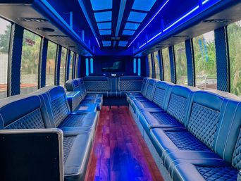 Vibrant blue-lit luxury party bus interior with quilted black leather bench seats along both sides, polished wood aisle, overhead skylights and panoramic windows showing palm trees outside.