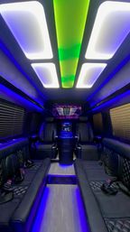 Luxury party van interior with black quilted leather seating, dual captain chairs, LED ceiling panels in green and white, blue ambient underlighting and wood-look floor.
