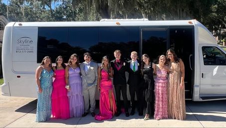 Prom-night group of friends in colorful formal gowns and tuxedos posing in front of a white party bus on a sunny suburban driveway