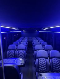 Sleek luxury shuttle interior with rows of quilted black leather captain seats, center aisle, wood-grain tables with cup holders, and cool blue LED ambient lighting