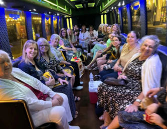 About twenty women smiling and chatting while seated facing each other inside a neon-lit party bus at night, bench seats, colorful LED strips, coolers and city lights visible through the windows.
