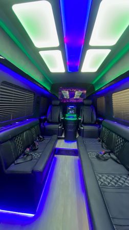 Luxury party bus interior with quilted black leather bench seating and captain chairs, wood-look aisle, and vibrant blue and green LED ceiling and floor lighting — party-ready vibe.
