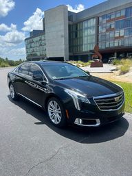 Black Cadillac luxury sedan parked on asphalt in front of a modern glass-and-concrete hotel entrance with a metal sculpture and blue sky