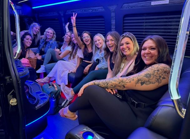 Smiling group of friends inside a neon-blue lit party bus, seated on leather benches with cupholders; one flashes a peace sign and another shows floral arm tattoos during a fun night out.