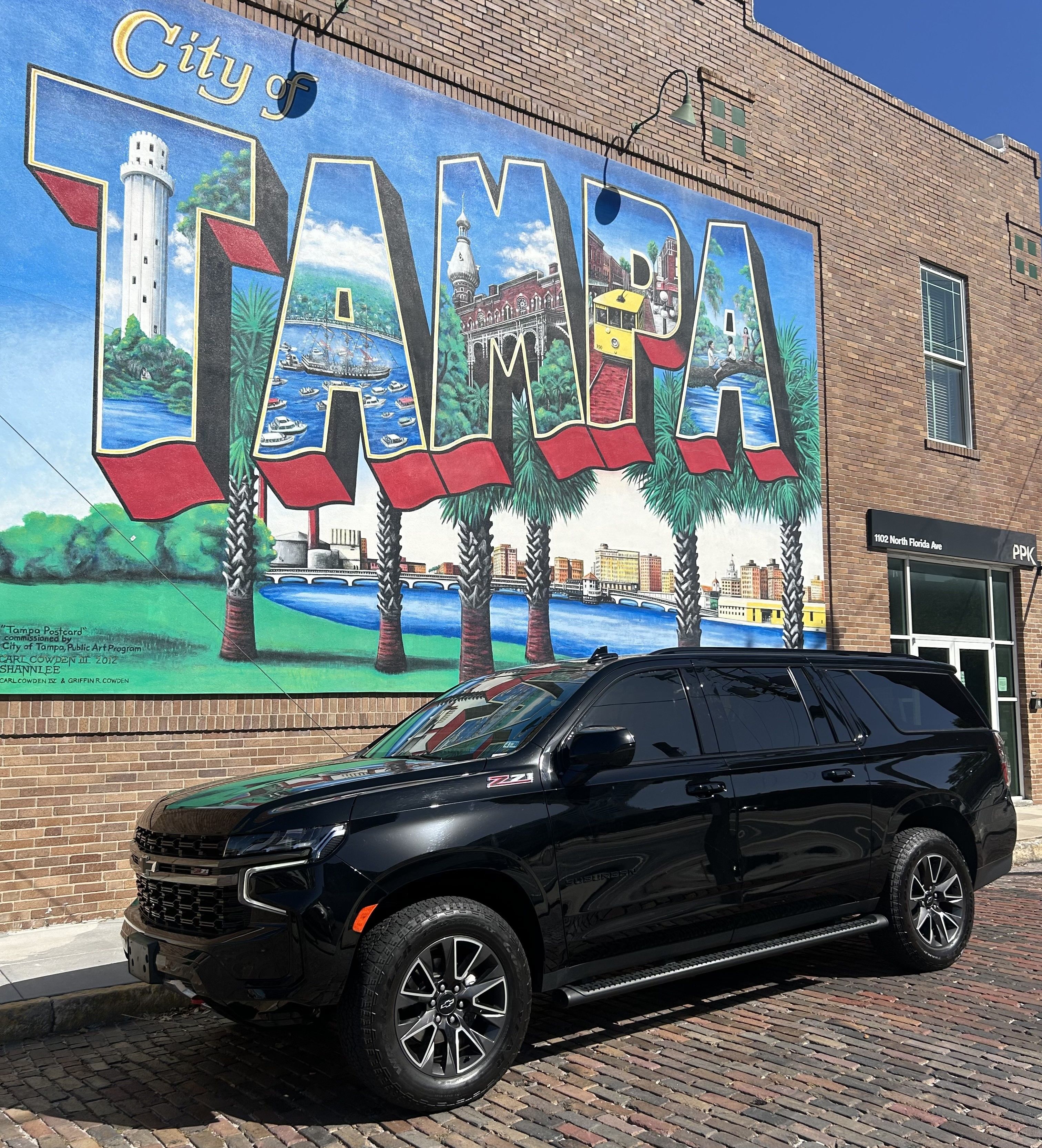 Colorful vintage-style "Tampa" mural on a brick building in Tampa, Florida, showing palm trees and waterfront scenes, with a black SUV parked on a cobblestone street beneath a blue sky.
