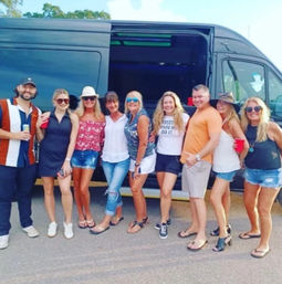 Nine adults in casual summer outfits smiling and posing with drinks in front of a black party van parked in a sunny outdoor lot — fun tailgate/road-trip gathering.