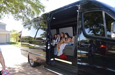 Group of teens in formal dresses and suits sitting inside a glossy black party van parked in a suburban driveway, trees and sunlight reflecting on the exterior — prom-night vibe.