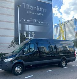 Black passenger shuttle van parked on a downtown street beneath a giant titanium phone billboard, with glass skyscrapers reflected in the van’s windows and blue sky overhead.
