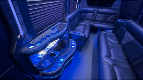 Blue LED-lit luxury party bus interior with black leather wraparound seating, illuminated cup holders and a curved wood-look drink console