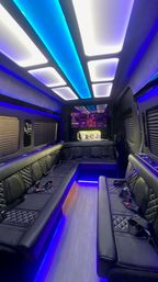 Sleek luxury party van interior with blue and white LED ceiling panels, U-shaped black quilted leather bench seating, seatbelts, wood-look floor and ambient under-seat lighting.