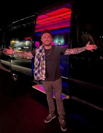 Smiling man with full-sleeve tattoos standing on a lit step with arms wide in front of a black party van glowing red and purple neon at night
