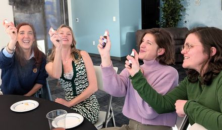 Four smiling women in a bright office meeting room, seated around a table with plates and cups, playfully spraying small sample bottles into the air.