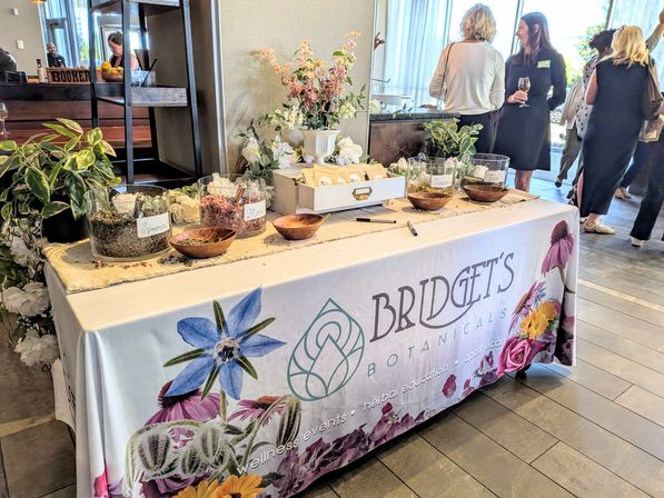 Indoor wellness pop-up vendor table with glass jars of herbs labeled spearmint, rose, chamomile and tulsi, wooden bowls, floral arrangements and people mingling nearby.