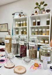 Herbal apothecary counter with labeled glass jars of dried herbs—Ginger, Lavender, Rose, Damiana and Tulsi—white shelving of more jars in the background and mixing tools, strainers and small beakers on the worktable.