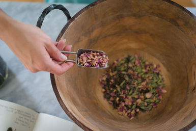 Hand holding a metal scoop of dried pink rose petals over a large wooden bowl filled with mixed dried petals and herbs, a DIY potpourri scene
