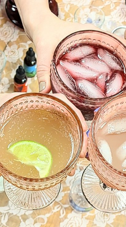 Overhead view of five hands toasting with vintage pink goblets of iced cocktails—assorted red, pink and citrus drinks with lime garnish—on a lace tablecloth at a casual cocktail party