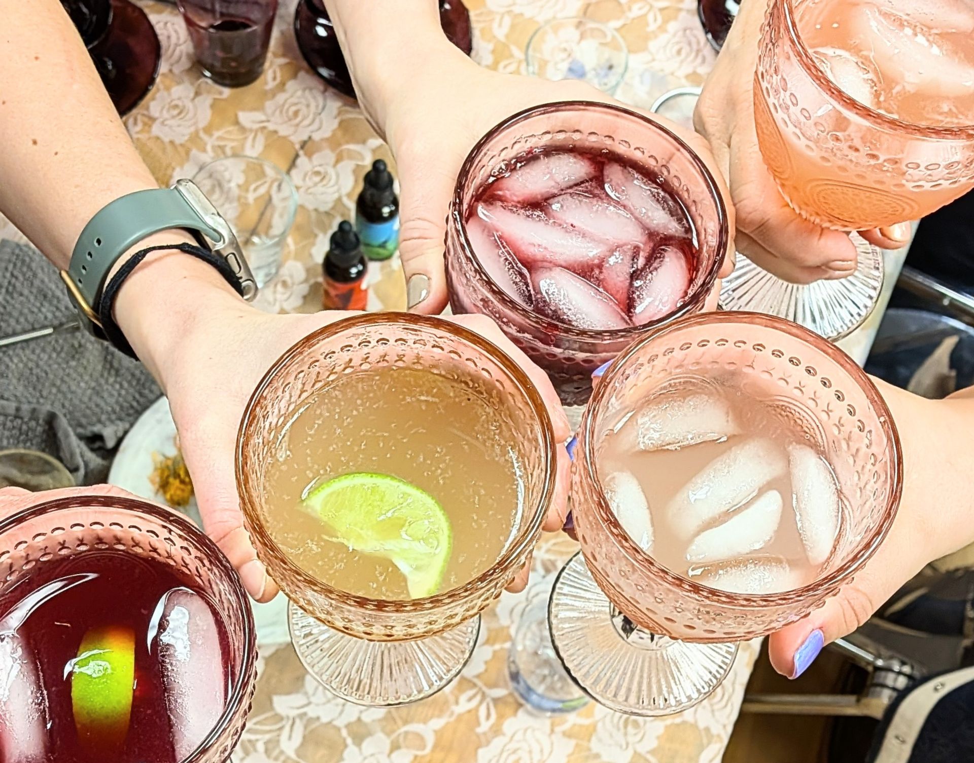 Overhead view of five hands toasting with vintage pink goblets of iced cocktails—assorted red, pink and citrus drinks with lime garnish—on a lace tablecloth at a casual cocktail party