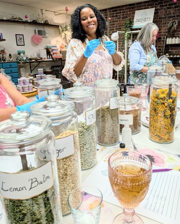 Smiling woman in an apron and blue gloves holding a small jar at an indoor herbal apothecary workshop, surrounded by labeled glass jars of dried herbs and botanicals.