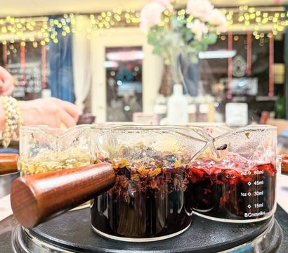 Three glass measuring pitchers with wooden handles steeping vibrant herbal brews—deep red hibiscus and pale chamomile—on a hot plate in a cozy tea bar with fairy lights bokeh.