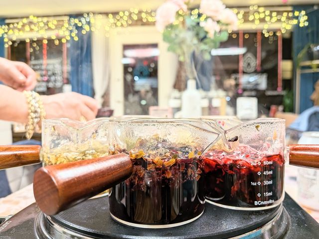 Three glass measuring pitchers with wooden handles steeping vibrant herbal brews—deep red hibiscus and pale chamomile—on a hot plate in a cozy tea bar with fairy lights bokeh.