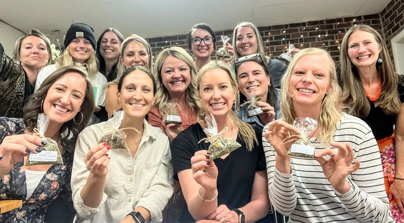 Smiling group of adult women at an indoor craft party holding small tied clear bags of dried herbs or tea as handmade gifts, posing in front of a brick wall with string lights.