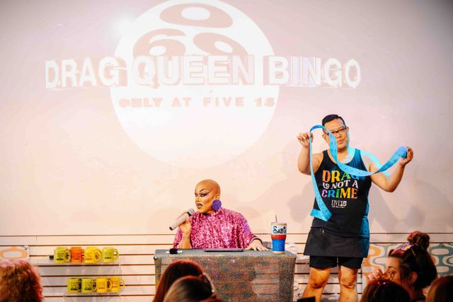 Drag Queen Bingo night: sequined host with microphone on stage and assistant unfurling blue bingo tickets under a projected 'Drag Queen Bingo' sign, colorful mugs on shelves and an engaged audience.