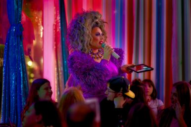 Drag performer in a glittery purple feathered outfit and big wig holding a sparkly microphone and tablet while hosting a lively cabaret-style drag show in a colorful neon nightclub with a seated audience.