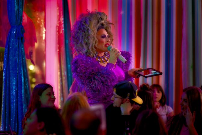 Drag performer in a glittery purple feathered outfit and big wig holding a sparkly microphone and tablet while hosting a lively cabaret-style drag show in a colorful neon nightclub with a seated audience.