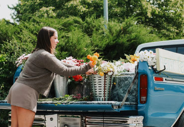 Woman arranging bright mixed flowers on the tailgate of a vintage blue pickup truck turned outdoor flower stand, parked in a leafy green setting.