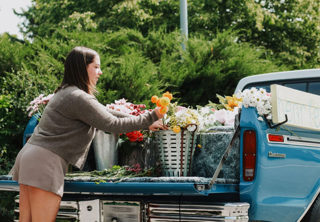 Woman arranging bright mixed flowers on the tailgate of a vintage blue pickup truck turned outdoor flower stand, parked in a leafy green setting.
