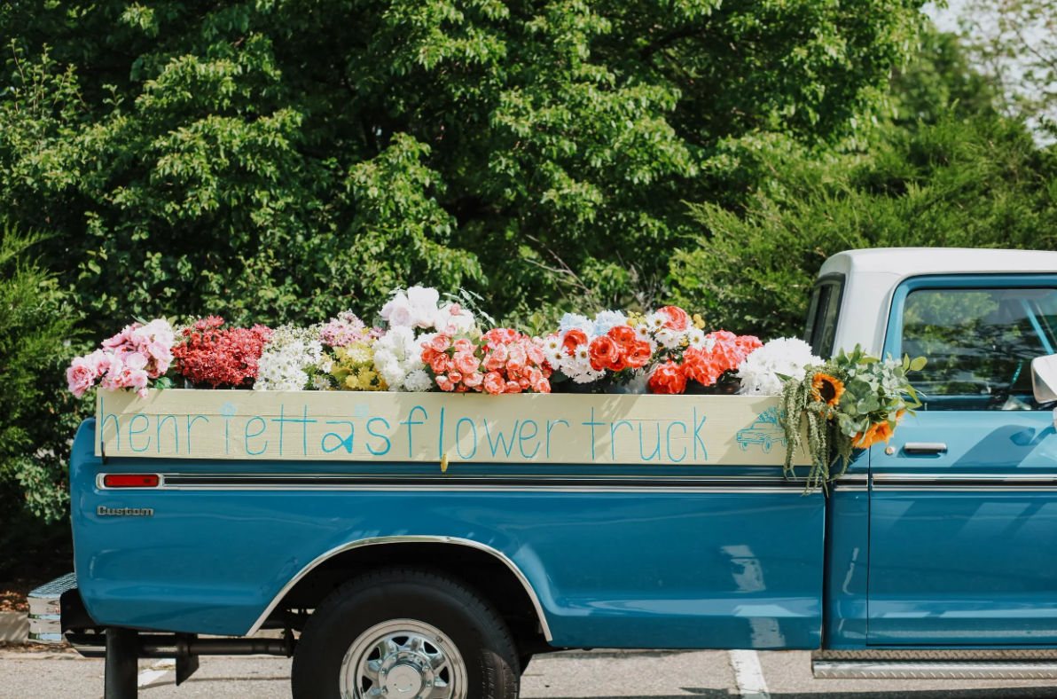 Cheerful blue vintage pickup truck parked by leafy greenery, bed overflowing with colorful bouquets and a hand-lettered flower-truck sign.