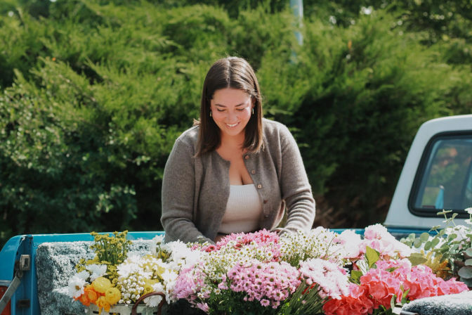 Smiling woman arranging pink, white and yellow bouquets in the bed of a blue pickup truck at a sunlit outdoor flower market, surrounded by lush greenery