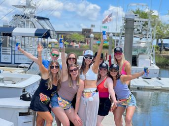 Eight women in summer outfits and sunglasses cheering with canned drinks on a marina dock in front of yachts, wearing colorful fanny packs and caps on a sunny day