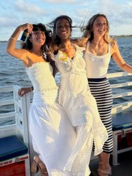 Three smiling friends in white summer outfits on a boat deck at golden hour, hair blowing in the breeze and one holding a canned drink, with calm water and a distant shoreline in the background.
