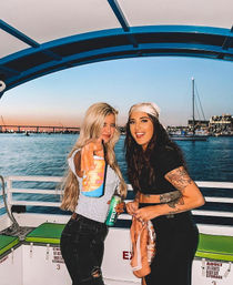 Two smiling women on a sunset boat cruise at a marina, holding canned drinks with sailboats and a waterfront skyline in the background.