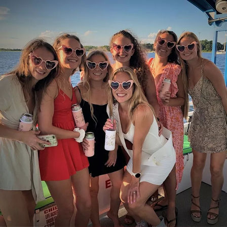 Seven friends in summer dresses wearing pink heart-shaped sunglasses, holding canned drinks on a boat at sunset over a lake