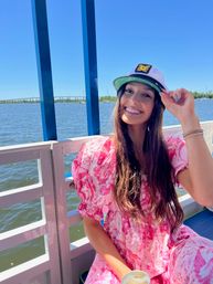 Smiling woman tipping a captain's hat on a sunny riverside boat ride, wearing a pink floral dress with blue water and a bridge in the background.