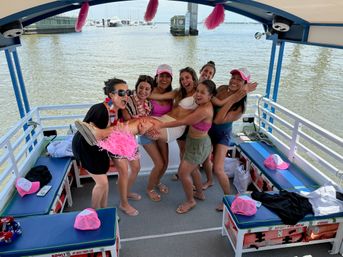 Seven friends laughing and lifting one friend on a pink-themed pontoon boat cruise near a marina, waterfront boat party with hats and tassel decorations.