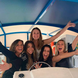 Five joyful friends cheering on a covered motorboat, gathered around the helm under a blue canopy at a marina