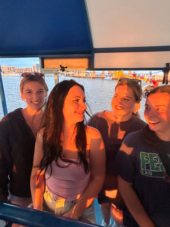 Four friends laughing on a harbor boat ride at sunset, warm golden light on their faces with marina docks and waterfront buildings in the background.