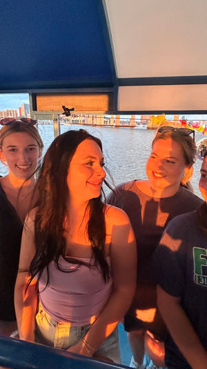 Four friends laughing on a harbor boat ride at sunset, warm golden light on their faces with marina docks and waterfront buildings in the background.
