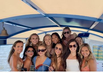 Group of smiling women in summer dresses and sunglasses gathered under a blue canopy on a boat, posing and enjoying a sunny lake cruise.